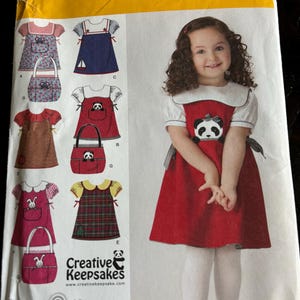 May include: A Simplicity sewing pattern for a girl's dress and matching bag. The pattern includes five different styles, including a red dress with a panda applique, a blue dress with a sailboat applique, and a plaid dress with a bunny applique. The pattern is for sizes 1/2, 1, 2, 3, and 4.