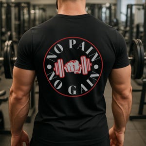 Op de afbeelding: Zwart T-shirt met een cirkelvormige afbeelding op de rug. De afbeelding toont de woorden "NO PAIN NO GAIN" in het wit, rondom een rode halter en een gebalde vuist. Het shirt heeft korte mouwen.