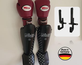 May include: Pair of burgundy boxing gloves and black shin guards with white patterned accents. The gloves and shin guards are hanging on a wall. Two black wall hooks are also visible, along with a "Made in Germany" sticker.