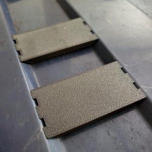 Könnte beinhalten: Zwei dunkelgraue rechteckige Objekte mit strukturierten Oberflächen und gekerbten Kanten. Die Objekte befinden sich auf einer dunkelblauen Oberfläche, wobei sich eines leicht über dem anderen befindet. Es handelt sich vermutlich um Komponenten oder Teile.