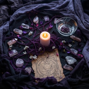 May include: A lit white candle surrounded by purple rope, crystals, and a vintage parchment with a star diagram. The arrangement includes a glass bowl and scattered flowers, all on a dark surface.