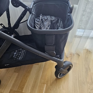May include: A black stroller with a dark interior and a black frame. The stroller has a front wheel with a white accent and is positioned on a light brown wooden floor.