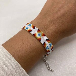 May include: A white beaded bracelet with a colorful geometric pattern of blue, orange, and red diamonds. The bracelet has a silver-colored chain clasp and is worn on a wrist.