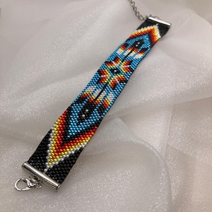 May include: A beaded bracelet featuring a geometric pattern in black, bright blue, yellow, orange, and white. The bracelet is finished with silver-toned metal end caps and a lobster claw clasp with an adjustable chain extension.