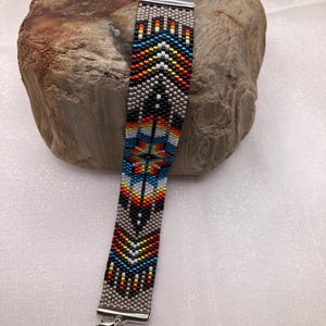 May include: A beaded bracelet with a Native American-inspired design. The bracelet features a pattern of black, gray, red, orange, yellow, and blue beads. It has a silver-tone clasp and chain for adjusting the length. The bracelet is approximately 1 inch wide.