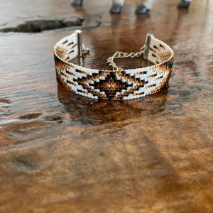 May include: A beaded bracelet with a geometric pattern in white, black, and brown. The bracelet has a silver-tone clasp and a chain extender. The bracelet is approximately 0.75 inches wide.