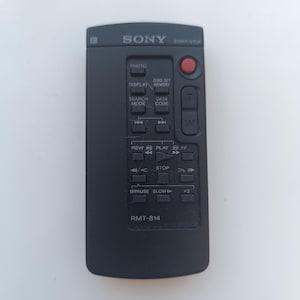 Pode incluir: Controle remoto preto para um gravador de vídeo Sony com botões para reproduzir, retroceder, avançar rápido, parar, pausar, diminuir a velocidade, foto, tela, modo de pesquisa, código de dados, memória de ajuste zero e iniciar/parar. O controle remoto tem o número do modelo RMT-814 impresso nele.