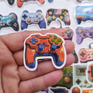 Pode incluir: Um autocolante de comando de videojogos laranja e azul é segurado numa mão, com uma coleção de outros autocolantes com tema de jogos no fundo. O autocolante tem botões verdes e uma borda branca. As palavras "GAME STORE" são visíveis.