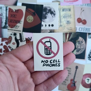Pode incluir: Um pequeno autocolante quadrado com um círculo vermelho e um ícone de telemóvel riscado. O texto "NO CELL PHONES" está em baixo. Outros cartões decorativos são visíveis no fundo.