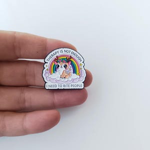Op de afbeelding: Een decoratieve emaille speld met een cartoonhert met gewei en een bloemenkrans, zittend op een wolk voor een regenboog. De tekst THERAPY IS NOT ENOUGH en I NEED TO BITE PEOPLE staat in zwarte hoofdletters op een witte achtergrond.