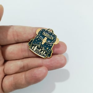 Könnte beinhalten: Ein dunkel-türkisfarbener Emaille-Pin in Rucksackform mit goldfarbenem Metallrand. Das Design zeigt ein Gebirge, Tannenbäume und Sterne, wobei das Wort TRAVELER in goldenen Großbuchstaben auf der oberen Klappe des Rucksacks abgebildet ist.