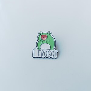 May include: A green enamel pin featuring a cartoon frog with a white belly and an open pink mouth. Below the frog, a white speech bubble shape contains the black text "I FROGOT" in a simple, rounded font, all outlined in a dark metal border.