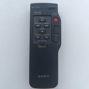 Pode incluir: Controle remoto preto para uma câmara de vídeo Sony Video 8. O controle remoto tem botões para reproduzir, retroceder, avançar rapidamente, parar, pausar, diminuir a velocidade e tela de dados. O controle remoto também tem um botão de retenção e um botão de iniciar/parar com um botão vermelho.
