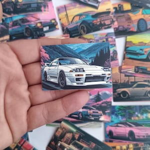 May include: A selection of stickers depicting illustrations of sports cars. The stickers display cars in bright colours, set against scenic backgrounds with mountains and cityscapes. The main sticker is a white sports car.
