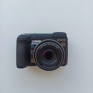 May include: A black digital camera with a silver lens. The lens has the text "KYOCERA 10X ZOOM LENS" and "f=5.7-57mm/2.8-3.1" printed on it. The camera has a red label that says "RTUNE features Fast".