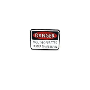 May include: A rectangular enamel pin featuring a white border and a black background. The top section contains a red oval with the word DANGER in white text. Below this, the text MOUTH OPERATES FASTER THAN BRAIN is printed in white.