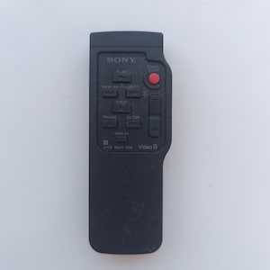 Pode incluir: Controle remoto preto para uma câmara de vídeo Sony Video 8. O controle remoto tem botões para reproduzir, retroceder, avançar rapidamente, parar, pausar, diminuir a velocidade e iniciar/parar. Também tem um botão de exibição e um rótulo de Video 8.