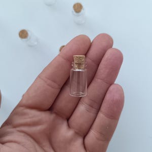 Puede incluir: Pequeñas botellas de vidrio transparente con tapones de corcho. Una botella se sostiene en una mano, con varias otras dispersas sobre una superficie blanca. Las botellas son cilíndricas y parecen estar vacías.