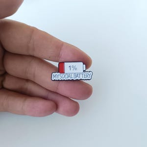 Op de afbeelding: Een kleine emaille speld in de vorm van een batterijpictogram met een rood gedeelte en de tekst 1% erin. Onder het batterijpictogram bevat een witte tekstballon de zwarte tekst MY SOCIAL BATTERY in een handgeschreven lettertype.