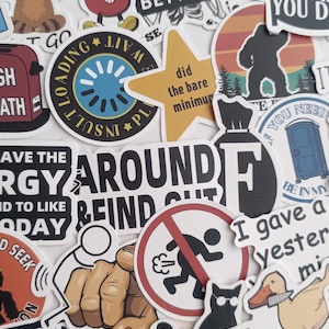 Op de afbeelding: Een verzameling diverse stickers met humoristische en motiverende ontwerpen. De stickers bevatten stripfiguren, zinnen als "Never Better" en illustraties van een luiaard, een kat en een eend met een mes. Het kleurenpalet omvat zwart, wit, rood en geel.