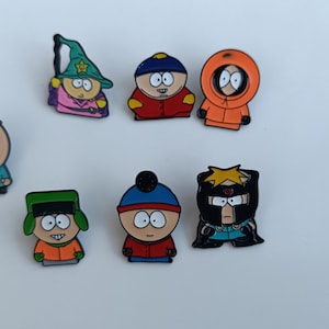 May include: A collection of seven colourful enamel pins featuring characters from the animated television series, South Park. The pins depict characters in their iconic outfits, including Cartman, Kenny, Stan, Kyle, and Butters.