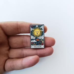 Könnte beinhalten: Eine rechteckige Emaille-Anstecknadel mit einem Tarotkarten-Motiv, das eine gelbe Sonne mit Gesicht, weiße Wolken und bunte Blumen auf dunkelblauem Grund zeigt. Oben steht die römische Ziffer X und unten der Schriftzug THE SUN.