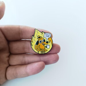 Può includere: Una spilla smaltata circolare raffigurante un cane giallo dei cartoni animati con un cappello, seduto su uno sgabello tra fiamme arancioni e gialle. Il cane tiene una tazza bianca e un fumetto contiene il testo nero THIS IS FINE.