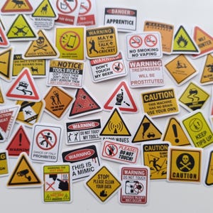 May include: A selection of hazard warning stickers in yellow, red, and white. The stickers display various symbols and text warnings, such as "Caution," "Warning," and "Danger." The stickers are arranged on a white surface.