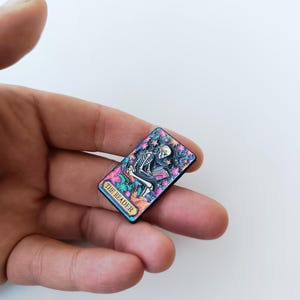 Peut inclure: Une broche émaillée rectangulaire représentant un squelette lisant un livre, entouré de fleurs et d'étoiles colorées. Le motif comprend une bannière dorée en bas avec le texte THE READER en lettres capitales noires sur fond sombre.