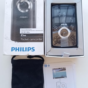 Pode incluir: Uma câmara de vídeo de bolso Philips ESee numa caixa branca com texto preto Full HD 1080p, uma bolsa preta com cordão e um manual de instruções. A câmara é preta com um botão de controlo circular prateado e um ecrã na parte frontal.