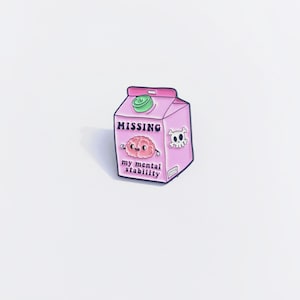 Op de afbeelding: Een roze emaille speld in de vorm van een melkpak met een cartoonillustratie van een brein. De tekst luidt MISSING en my mental stability. Een klein wit doodshoofd met gekruiste botten staat aan de zijkant en een groene dop zit bovenop.