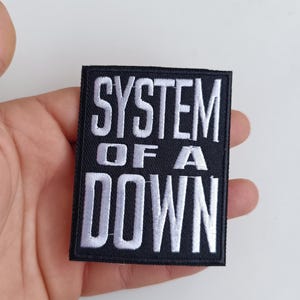 Może przedstawiać: Czarna kwadratowa naszywka z białym haftowanym napisem „SYSTEM OF A DOWN”. Naszywka zawiera nazwę zespołu w odważnej, stylizowanej czcionce. Naszywka jest prawdopodobnie przeznaczona na odzież lub akcesoria.