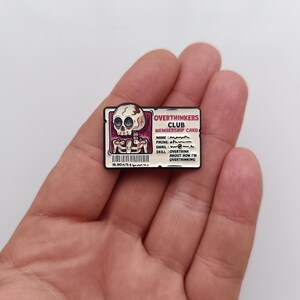 May include: A novelty pin with a skull design and the words "OVERTHINKERS CLUB MEMBERSHIP CARD". The card includes a barcode and details such as name, phone, email, and skill: overthink. The pin is held in a hand.