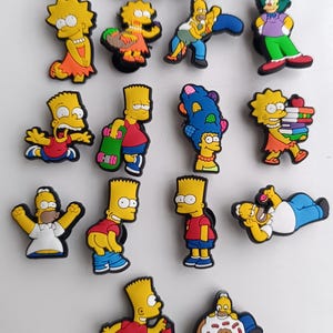 May include: A selection of colourful cartoon character charms, featuring characters from The Simpsons. The charms depict various characters in different poses and outfits, including Homer, Bart, Lisa, and others. The charms are made of a glossy material.