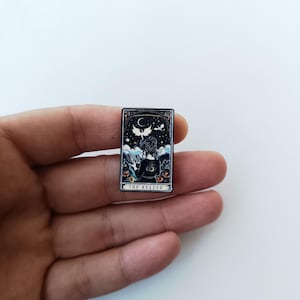 Pode incluir: Um broche de esmalte retangular com uma ilustração escura de uma pessoa lendo um livro sob um céu noturno com uma lua crescente e um dragão voador. O design inclui flores laranja e o texto THE READER na parte inferior.