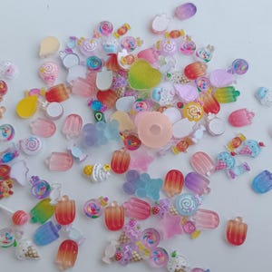 Könnte beinhalten: Eine Sammlung bunter, miniaturhafter Harzanhänger in verschiedenen Formen, darunter Lutscher, Eiswaffeln und Süßigkeiten. Die Anhänger sind in Rosa-, Lila-, Gelb- und Blautönen gehalten und auf einer weißen Oberfläche verstreut. Ideal für Bastelarbeiten und Schmuckherstellung.