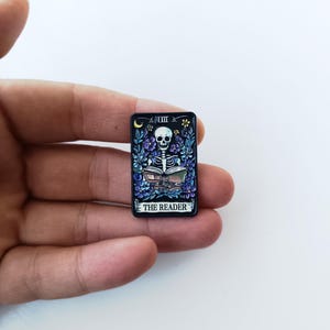 Pode incluir: Um broche de esmalte retangular com fundo preto apresentando um esqueleto branco lendo um livro aberto. O design inclui flores azuis e roxas, uma pequena lua crescente amarela e o texto THE READER em letras brancas.