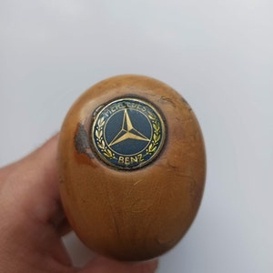 Op de afbeelding: Een houten versnellingspookknop met een gouden Mercedes-Benz-logo. Het logo heeft een driepuntige ster en de tekst "Mercedes-Benz".