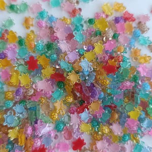Può includere: Una collezione di abbellimenti traslucidi a forma di tartaruga in varie tonalità di rosa, giallo, verde, blu e arancione. Questi piccoli pezzi decorativi sono sparsi su una superficie bianca, creando un'esposizione giocosa e vibrante.