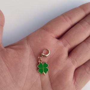 Pode incluir: Um berloque dourado com um design de trevo de quatro folhas verde. O berloque tem um pequeno fecho para prender a uma pulseira ou colar. O trevo é contornado em dourado.