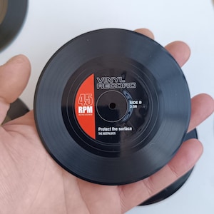 Puede incluir: Posavasos de disco de vinilo negro con texto blanco y rojo. El posavasos dice "VINYL RECORD" y "45 RPM". También es visible el texto "Protect the surface". La cara B es 3:56.
