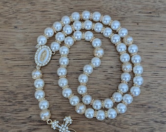 Può includere: Un rosario con perle bianche finte alternate a distanziatori in metallo dorato. Il pezzo include un ciondolo ovale raffigurante una figura in rilievo circondata da piccole pietre chiare e una croce tempestata di pietre trasparenti.