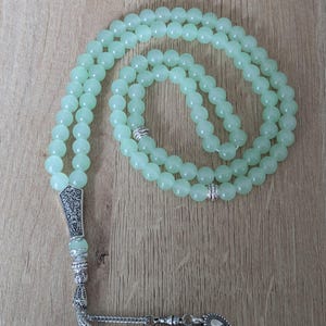 May include: A light green beaded prayer beads, or tasbih, with a decorative silver-colored pendant and tassel. The beads are round and evenly sized. The background is a light brown wooden surface.