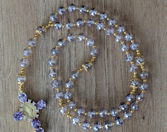 May include: A rosary with faceted, light purple beads and gold-colored accents. The cross pendant is adorned with purple gemstones and a central medallion. The rosary is displayed on a wooden surface.