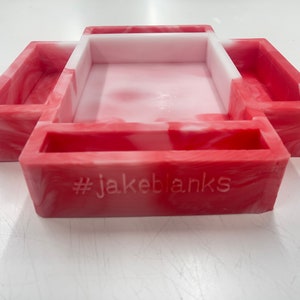 May include: A red and white marbled resin tray with three compartments. The tray has a rectangular central section and two side sections. The text "#jakeblanks" is engraved on the front of the tray.