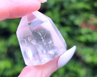 Puede incluir: Una punta de cristal de cuarzo transparente con una parte superior facetada y una base natural rugosa. El cristal se sostiene contra un fondo verde borroso, mostrando su transparencia y sutil tono rosado. Este cristal es una piedra preciosa natural.