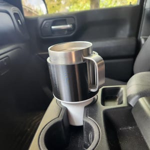 May include: A stainless steel travel mug with a black and white cup holder, placed in a car's center console. The mug has a silver handle and a black exterior. The cup holder is designed to fit the car's cup holder.