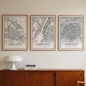 May include: Three vertical framed wall art prints featuring black and white street map designs. Each print displays a detailed city grid, the text YOUR CITY, YOUR COUNTRY, and geographic coordinates at the bottom, all encased in light wood frames.