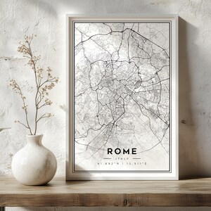 May include: A framed wall art print featuring a black and white street map of Rome, Italy. The print includes the text ROME, ITALY, and the coordinates 41.892 N | 12.511 E centred at the bottom within a thin black rectangular border.