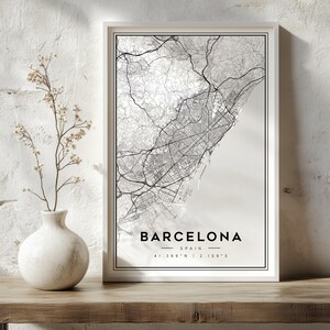 May include: A framed vertical wall art print featuring a black and white map of Barcelona, Spain. The print displays intricate street grids and topography, with the text BARCELONA, SPAIN, and the coordinates 41.389 N | 2.159 E printed below.